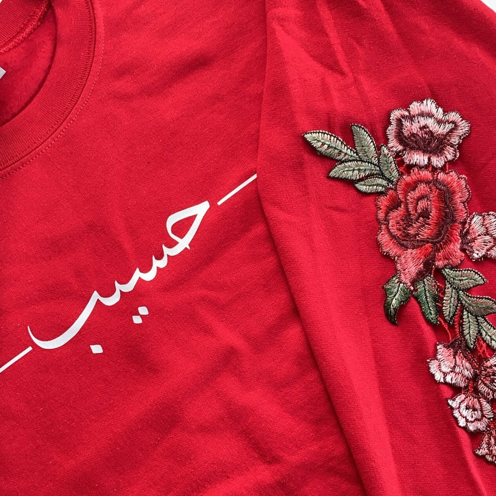 red sweatshirt with white Arabic name in font 3 printed across the chest with 2 straight horizontal lines and red sleeve embroidery