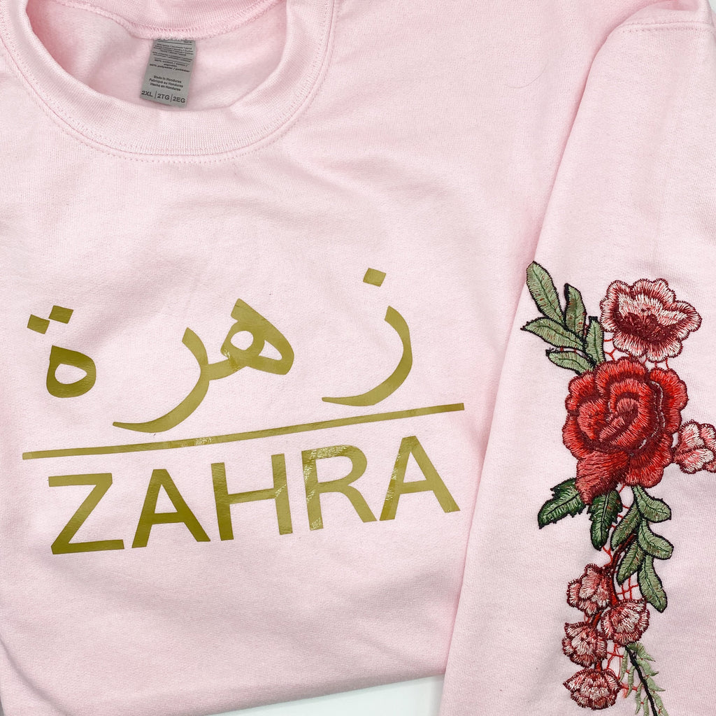 light pink sweatshirt with metallic gold design Arabic and English name and red floral sleeve embroidery