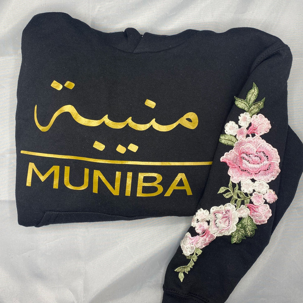 black hoodie with metallic gold Arabic and English name across chest with pink floral sleeve embroidery