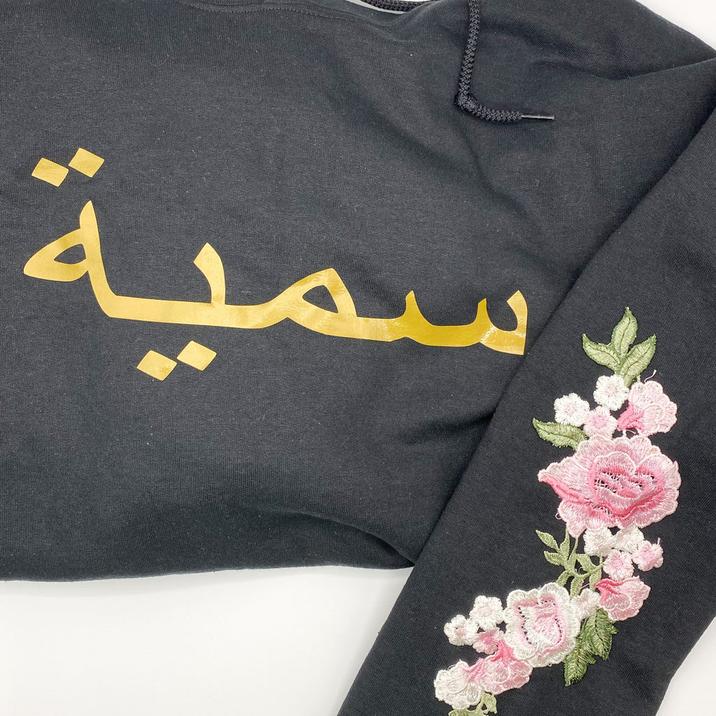 black sweatshirt with metallic gold Arabic name and pink floral sleeve embroidery