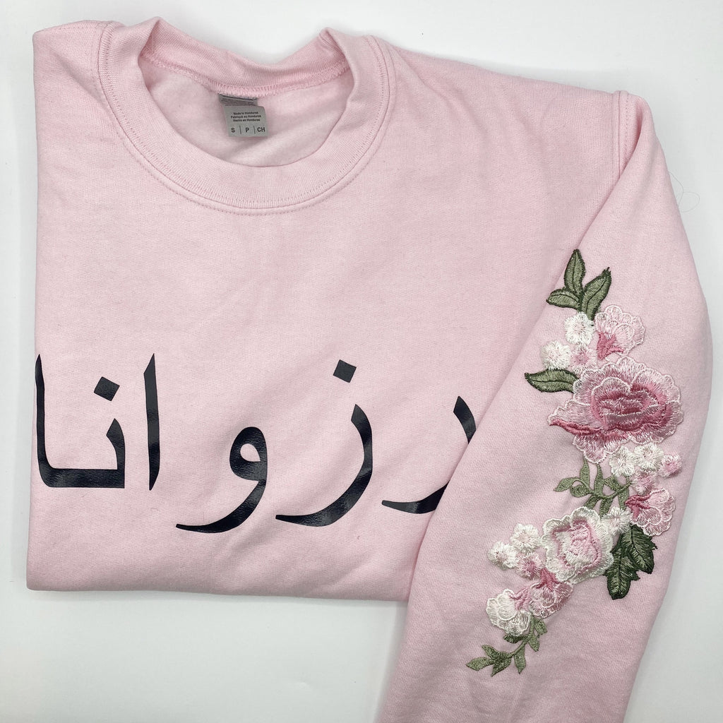 light pink sweatshirt with black Arabic name across chest and pink floral sleeve embroidery