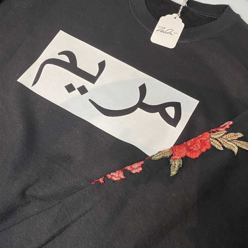 black sweatshirt with white box and black Arabic name and red floral sleeve embroidery