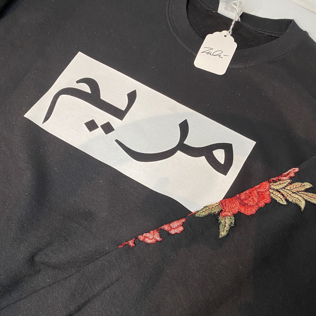 black hoodie with white box and black Arabic name across chest and red floral sleeve embroidery