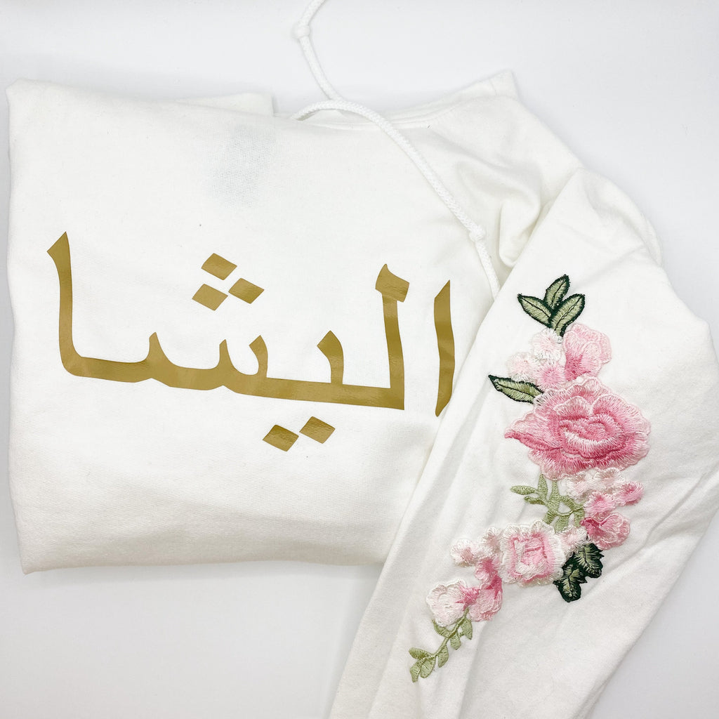 white sweatshirt with metallic gold Arabic name and pink floral sleeve embroidery
