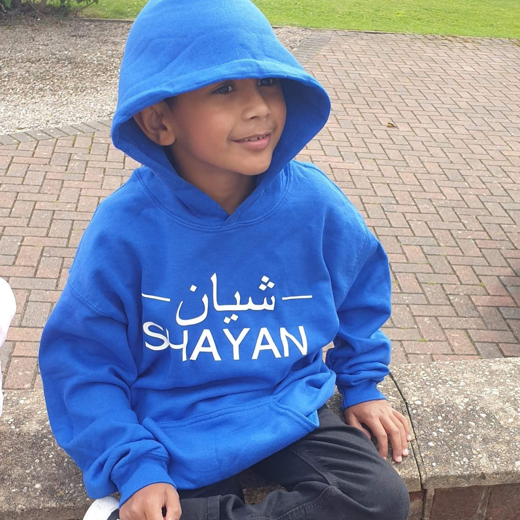 boy wearing royal blue hoodie with white design printed across the chest. design includes Arabic name in the middle with 2 lines on either side and English name underneath