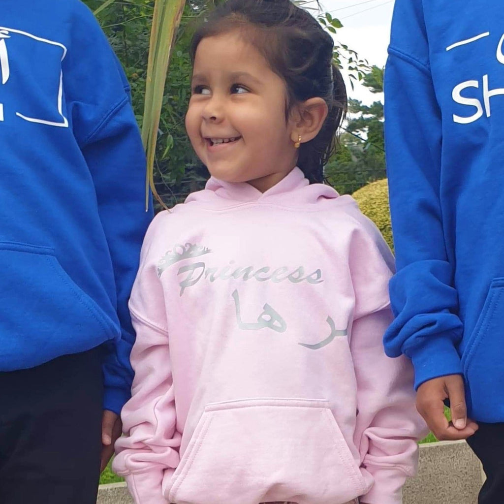 a girl wearing a personalised princess hoodie. Girl is wearing a baby pink hoodie with silver design. Design includes a tiara with 'princess' and her name in Arabic