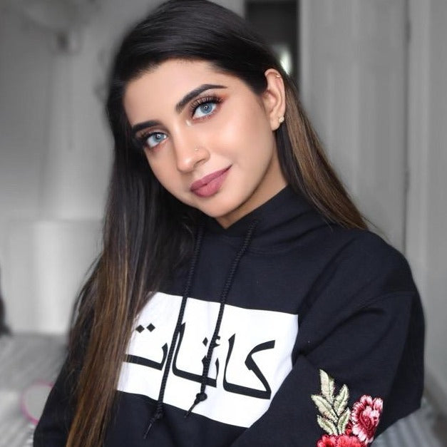 Model wearing black hoodie with white rectangle across chest and black Arabic name inside rectangle and red floral sleeve embroidery