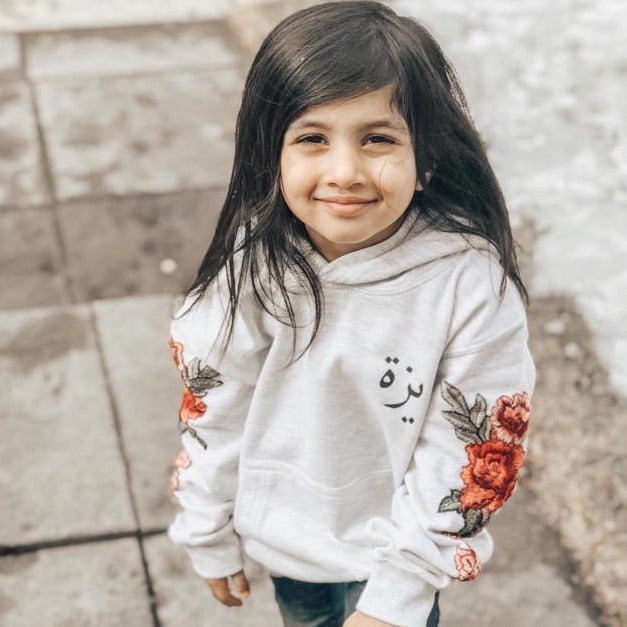 Customer Photo - girl wearing ash grey hoodie with black arabic name on left chest (font 2) and red floral sleeve embroidery
