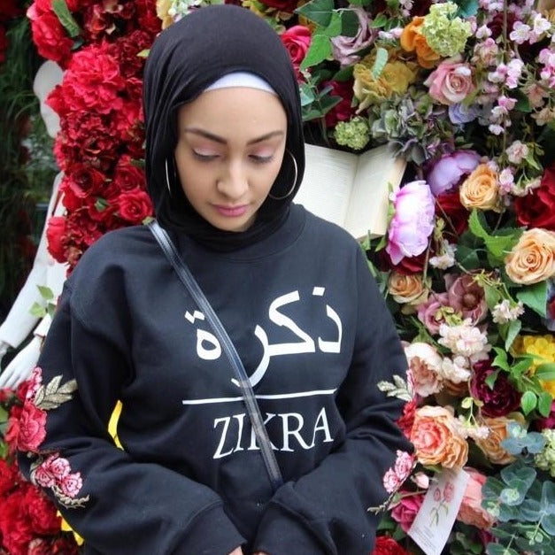 model wearing black sweatshirt with white Arabic font 1 and English name across the chest and red sleeve embroidery